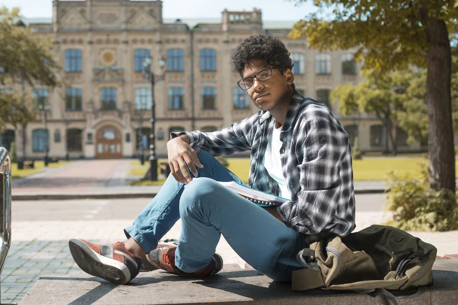 Black male student in glasses sitting on a low wall outside a university building, holding a notebook, with a backpack beside him